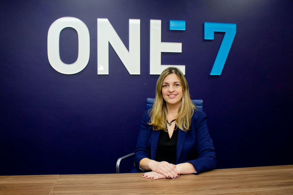 Patricia Baceti sentada com as maos em uma mesa e o logo da One7 ao fundo Patricia Baceti sentada com as maos em uma mesa e o logo da One7 ao fundo
